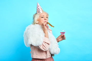 Elderly beautiful woman blowing a party horn, holding a glass of juice, against blue...