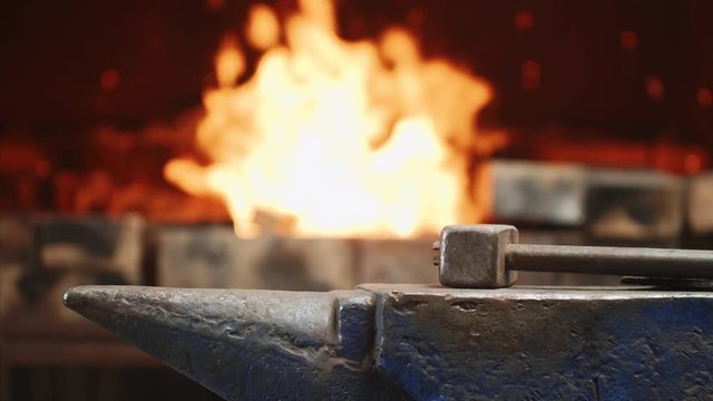 Workplace for forging metal. Hammer on anvil in forge on fire background. Worker throws garbage in furnace from shovel, back view. Smithy workshop in the metallurgical industry. Incineration of waste.