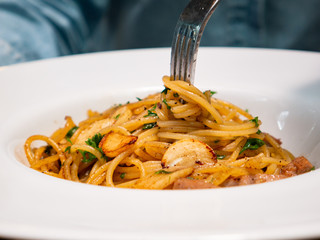A female hand is trying to use fork to eat Spicy spaghetti with pork sausage and garlic in white bowl, Italian cuisine