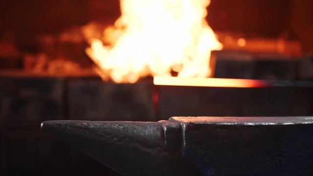 Smith takes off hot metal bar from furnace fire and puts on anvil. Blacksmith is hitting hot metal bar with a massive hammer on anvil, hands closeup. He is working in smithy workshop in slow motion.