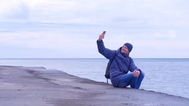 Tourist in warm clother hat and jacket takes a pictures on mobile phone on sea background. Man traveller makes selfie on smartphone aits on waterfront at sea in winter. Overcast rainy day at seaside.