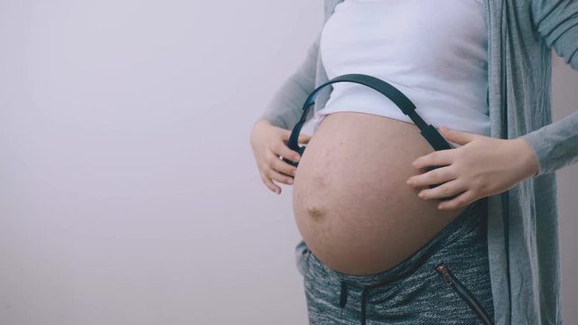 young pregnant lady in grey jacket puts modern headphones on large bare belly standing at white wall closeup