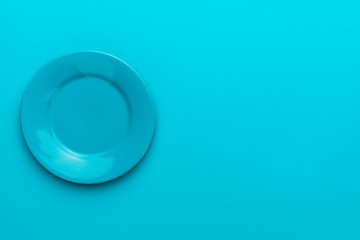 Image of round ceramic plate over turquoise blue background with copy space and left side position. Top view of blank dish on blue table.