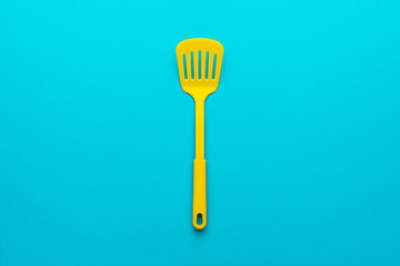 Top view of plastic kitchen utensil. Minimalistic flat lay image of yellow slotted spatula over turquoise blue background with copy space. Central composition of turning spatula on blue backdrop.