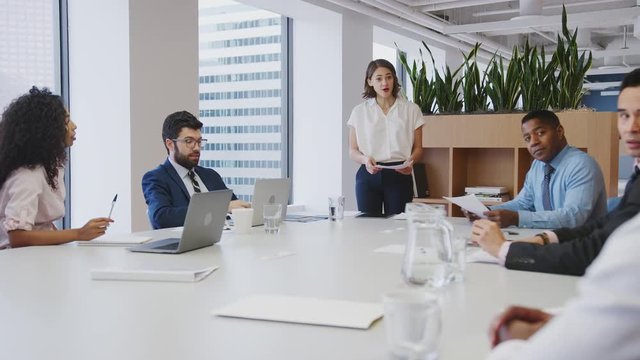 Businesswoman Standing Giving Presentation To Colleagues Sitting Around Table In Modern Office 