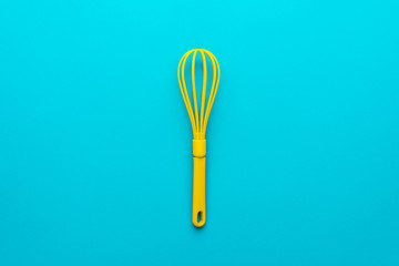 Top view photo of vivid plastic kitchen utensil. Minimalist flat lay image of yellow whisk over turquoise blue background with copy space and central composition. Mixing kitchen tool on blue table.