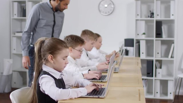 Medium arc shot of preteen primary school pupils sitting in classroom at computer education class and learning to type on laptops, while African American male teacher is checking work and helping