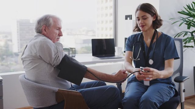 Nurse Wearing Scrubs In Hospital Office Checking Senior Male Patients Blood Pressure