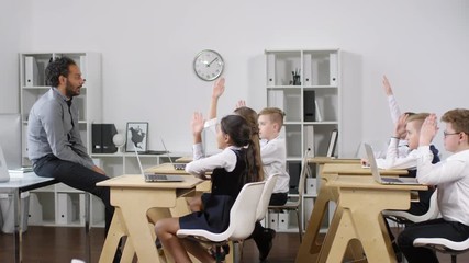 Full side shot of enthusiastic Afro-American male teacher sitting on table during e-learning class and asking questions to group of primary school pupils, who are eagerly putting up hands to answer
