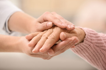 Nurse holding hands of elderly woman against blurred background, closeup. Assisting senior generation