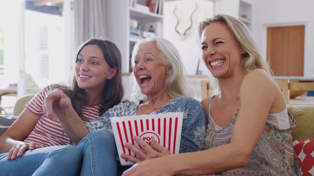 Mother With Adult Daughter And Teenage Granddaughter Eating Popcorn Watching Movie On Sofa At Home 