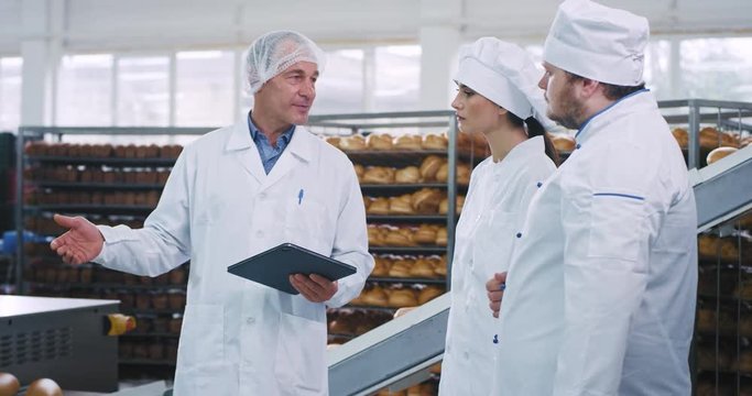Meeting time at bakery factory old man engineer with a performance tablet explaining the rules of using the industrial machine to bakers pretty lady and man