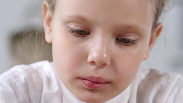 Close-up face shot of young Caucasian schoolgirl with chapped lips, wearing white blouse, looking down at something then sighing