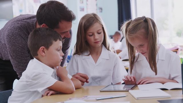 Male Teacher With Three Elementary School Pupils Wearing Uniform Using Digital Tablet At Desk