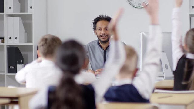 Medium shot of cheerful young black teacher with curly Afro hair giving lesson for primary school pupils and asking them questions, and children eagerly raising their hands to answer