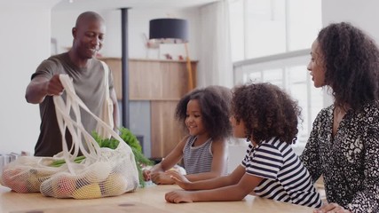 Father Returns Home From Shopping Trip As Mother Helps Children With Homework On Kitchen Table