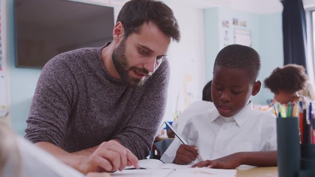 Elementary School Teacher Giving Male Pupil Wearing Uniform One To One Support In Classroom 
