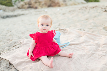 Cute baby girl in a red dress sitting and playing on the beach with sand