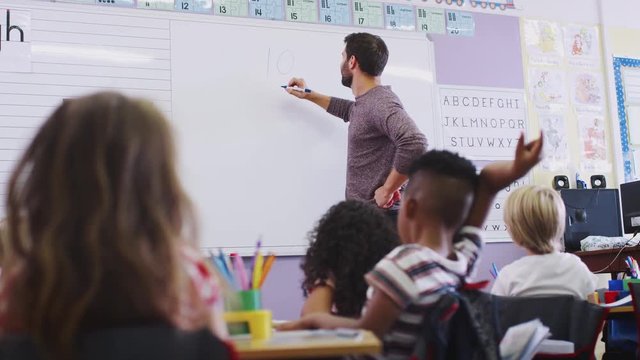 Male Teacher Standing At Whiteboard Teaching Lesson To Elementary Pupils In School Classroom