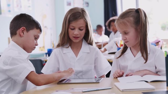 Three Elementary School Pupils Wearing Uniform Using Digital Tablet At Desk