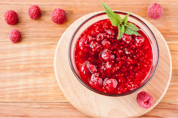 Glass small saucer with raspberry jam with fresh raspberries and mint close-up on wooden background.Copy space