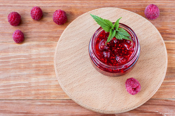 Glass small jar with raspberry jam with fresh raspberries and mint close-up on wooden background.Copy space
