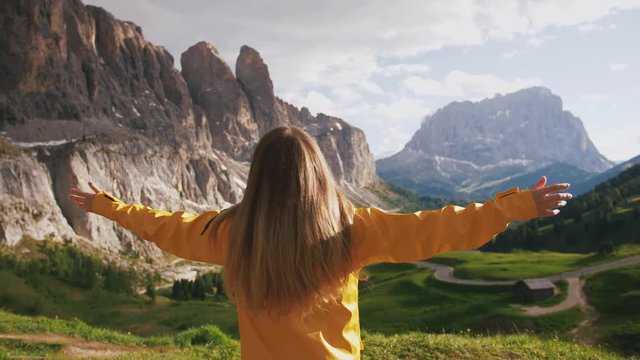 Young cheerful woman in a yellow raincoat rising hands and enjoying view of beautiful mountains, slow motion