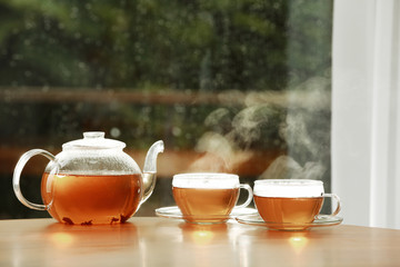 Teapot and cups of hot tea on wooden table against blurred background, space for text