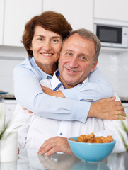 Mature family couple hugging and sitting at kitchen table while drinking tea