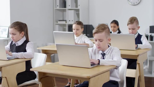 Panning medium shot of five preteen schoolchildren, wearing school uniform, sitting at desks in brightly lit classroom and all diligently typing on laptops