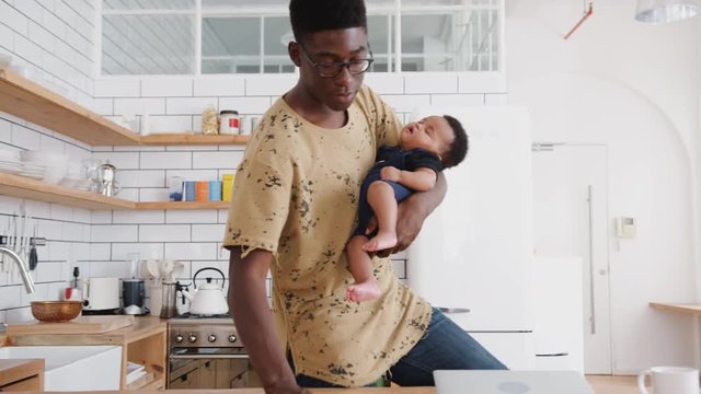 Multi-Tasking Father Holds Sleeping Baby Son Whilst Cleaning And Working On Laptop In Kitchen
