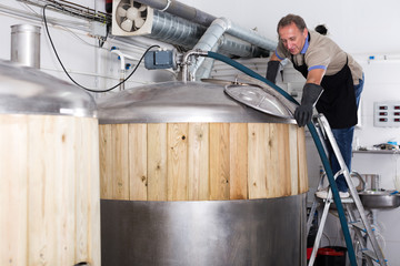 Adult brewer is standing with tube near reservoir for beer