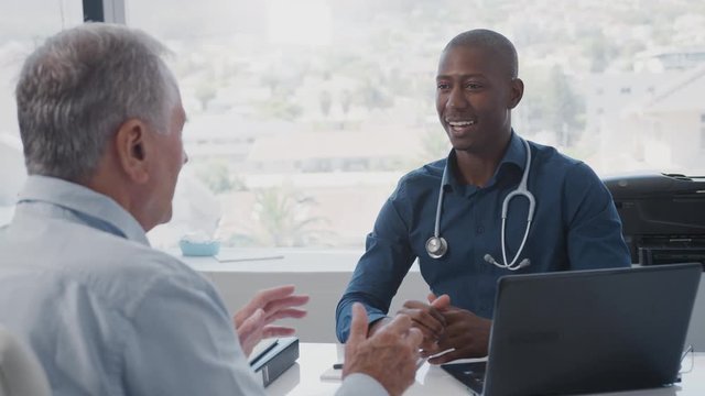 Senior Male Patient In Consultation Shaking Hands With Doctor Sitting At Desk In Office
