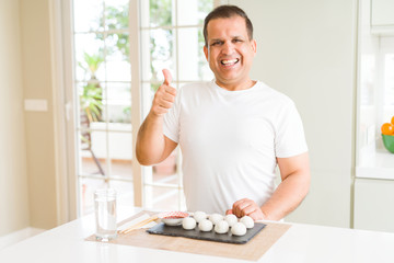 Middle age man eating asian dumplings at home doing happy thumbs up gesture with hand. Approving expression looking at the camera with showing success.