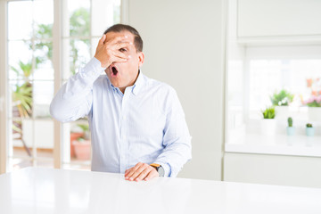 Fototapeta premium Middle age man sitting at home peeking in shock covering face and eyes with hand, looking through fingers with embarrassed expression.