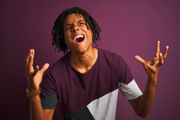 Afro man with dreadlocks wearing casual t-shirt standing over isolated purple background crazy and mad shouting and yelling with aggressive expression and arms raised. Frustration concept.