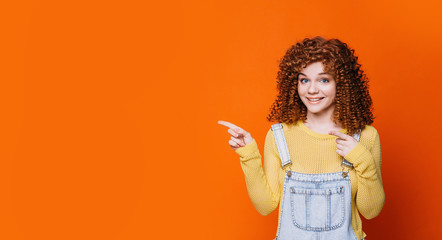 red-haired woman with curly hair advertising product, pointing hands on empty orange background