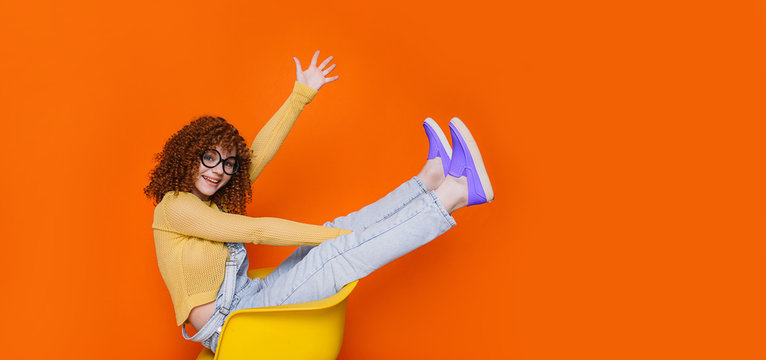 Happy emotionally trendy teenage sitting on chair with raised legs, wearing stylish violet slipoon