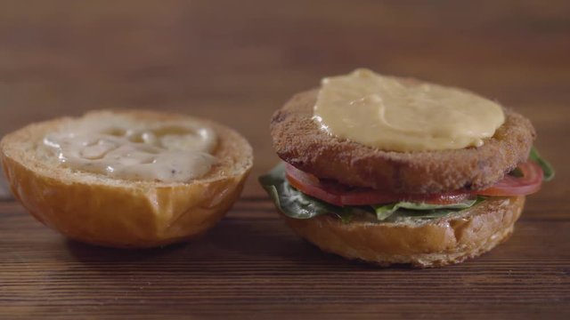 Hands of the chef making burger close-up. The cook putting melted cheese on the cutlet, tomato slices and basil leaves lying on the one piece of a hamburger bun. Tasty food preparation