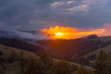 Bright sunset over foggy mountains. Vivid sun through the clouds over mountains slopes, covered with spruce forest. Carpathian mountains. Ukraine.