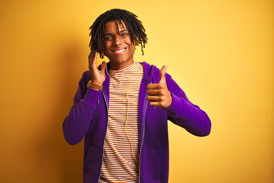 Afro man with dreadlocks listening to music usig headphones over isolated yellow background happy with big smile doing ok sign, thumb up with fingers, excellent sign