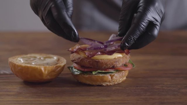 Hands of the chef making burger close-up. The cook putting becon and onion on the cutlet, tomato slices and basil leaves lying on the one piece of a hamburger bun. Tasty food preparation