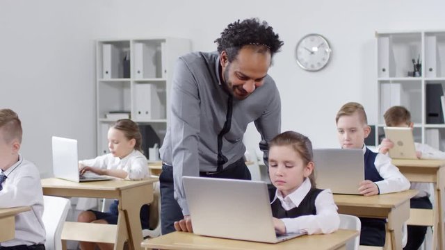 Tracking shot of elementary school pupils sitting at desks with laptops during ICT class and working on assignment, and charismatic African American teacher standing next to schoolgirl and explaining