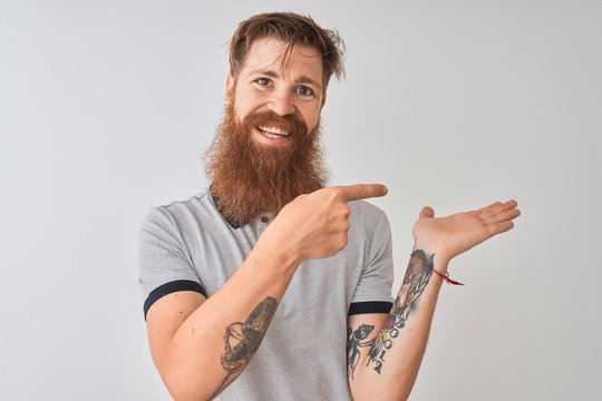 Young redhead irish man wearing grey polo standing over isolated white background amazed and smiling to the camera while presenting with hand and pointing with finger.