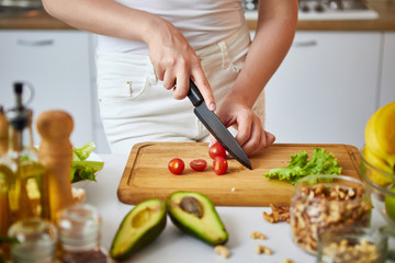 Young happy woman cutting tomatoes for making salad in the beautiful kitchen with green fresh ingredients indoors. Healthy food and Dieting concept. Loosing Weight