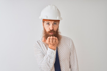 Young redhead irish architect man wearing security helmet over isolated white background looking at the camera blowing a kiss with hand on air being lovely and sexy. Love expression.