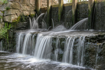A small waterfall near a water mill shot with a long exposure and blurred water, like milk. Water flows through the old stonework. Old wet stones from which grass grows.