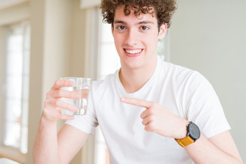 Young man drinking a glass of water at home very happy pointing with hand and finger