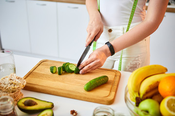Young happy woman cutting cucumber for making salad in the beautiful kitchen with green fresh ingredients indoors. Healthy food and Dieting concept. Loosing Weight