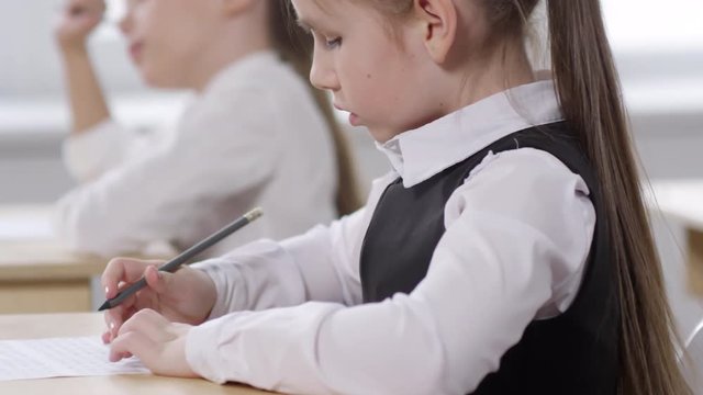 Tracking close-up shot of Caucasian preteen schoolgirl, dressed in uniform white shirt and pinafore, sitting at desk in classroom, writing in test answer sheet and asking classmate for help